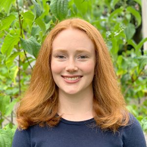 Sarah Gediman's headshot depicts her smiling, standing in front of a green shrub.