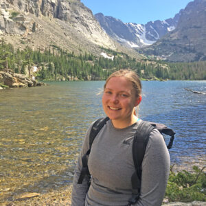 Smiling photo of Annali Cers standing in front of a body of water framed by mountains.